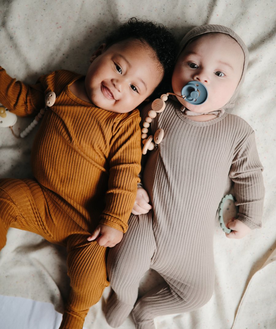 Dois bebés deitados com macacões de malha canelada, um castanho alaranjado e outro bege, um com chucha azul.