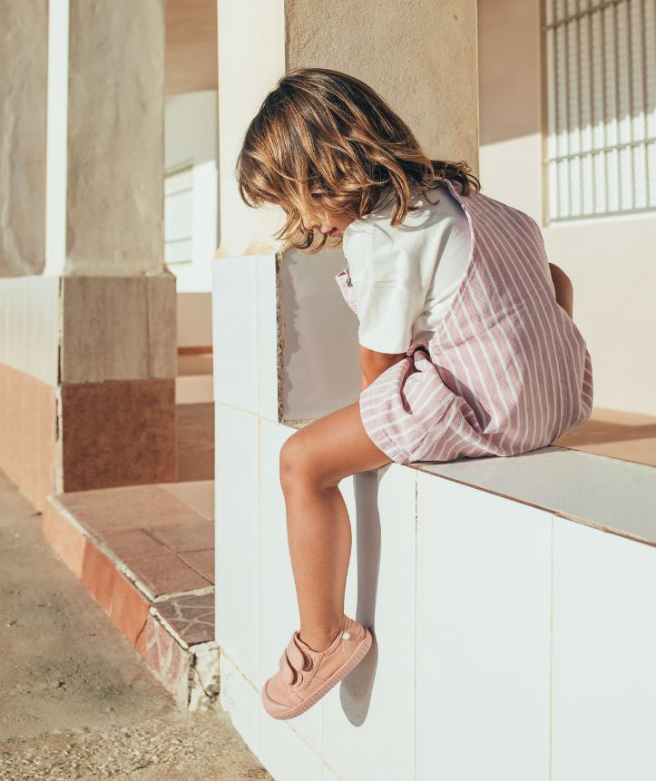 Criança sentada de vestido rosa às riscas numa parede branca com ténis cor de rosa.