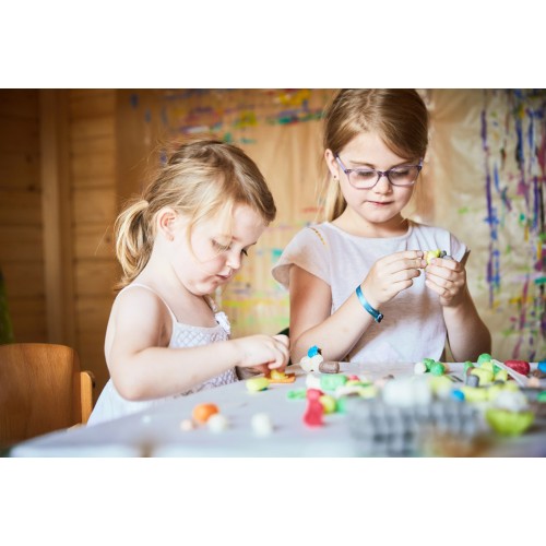Duas crianças a brincar com pequenos brinquedos coloridos numa mesa branca num quarto com paredes de madeira