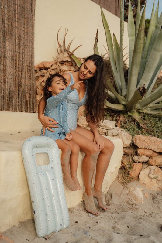 Mulher e criança sentadas junto a colchão insuflável azul em ambiente de praia