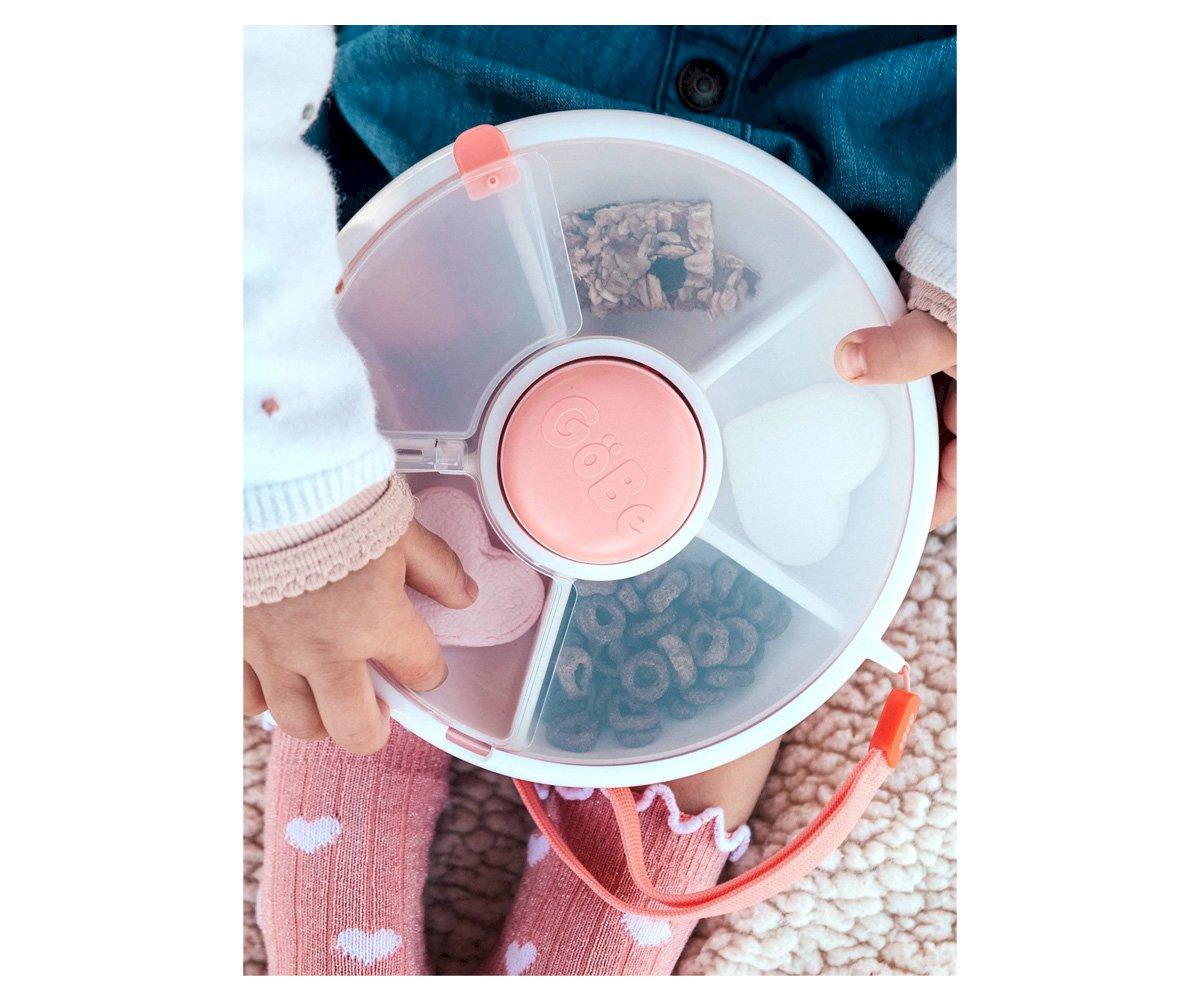 Recipiente de plástico dividido em cinco compartimentos com tampa rosa, contendo cereais, nas mãos de uma criança com meias rosa com corações brancos