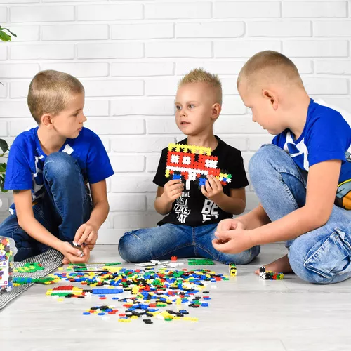 Três crianças brincando com peças de LEGO coloridas no chão branco junto a parede de tijolos branca.