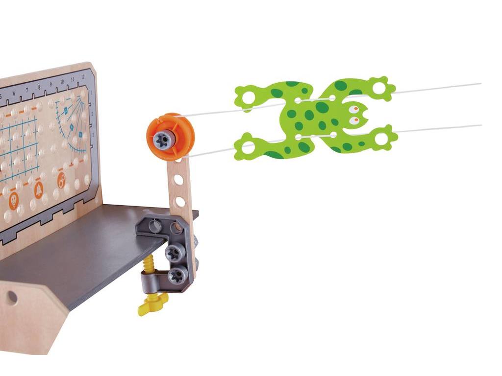 Dispositivo de madeira com rã verde pendurada em fios elásticos, montado numa superfície clara.