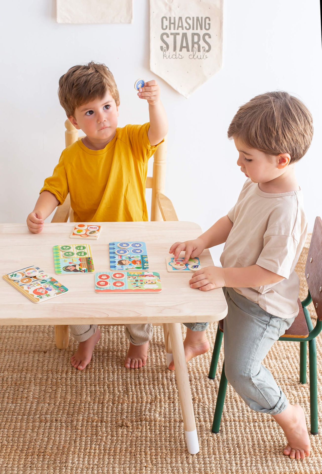 Crianças jogando jogo de tabuleiro com cartas e fichas à mesa de madeira, pano na parede com texto CHASING STARS kids club.
