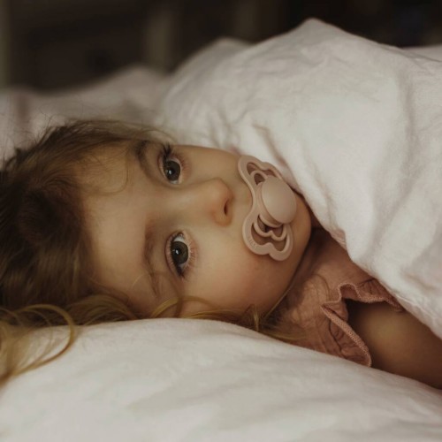 Bebé deitada com chupeta rosa claro e lençóis brancos numa cama