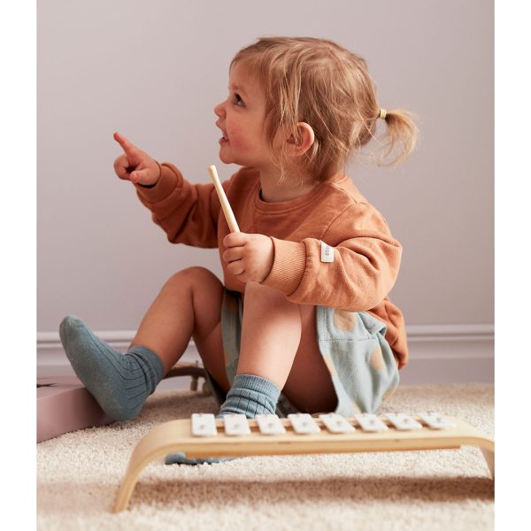 Criança sentada com roupa castanha e azul ao lado de um xilofone de madeira