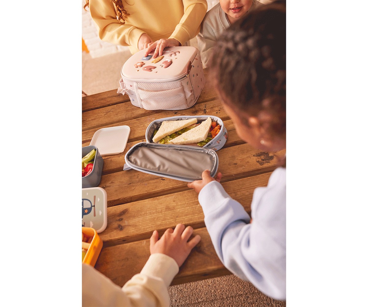 Crianças em volta de mesa de madeira com lancheira rosa e recipientes de comida.