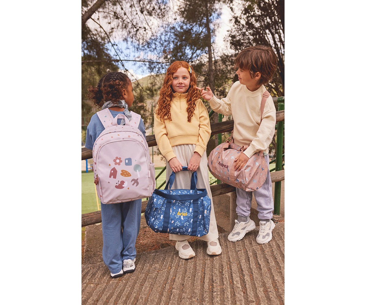 Três crianças com mochilas e malas de diferentes cores e padrões, ao ar livre