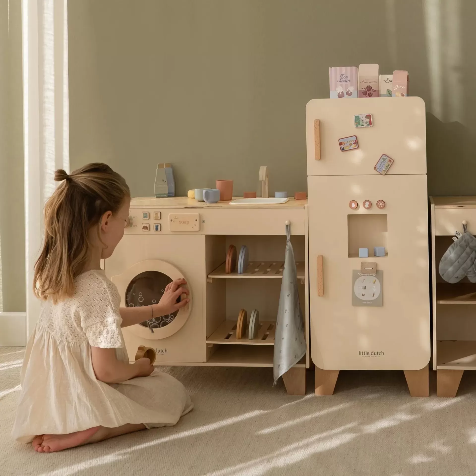 Criança brinca com brinquedo de cozinha em madeira creme com frigorífico e máquina de lavar roupa.