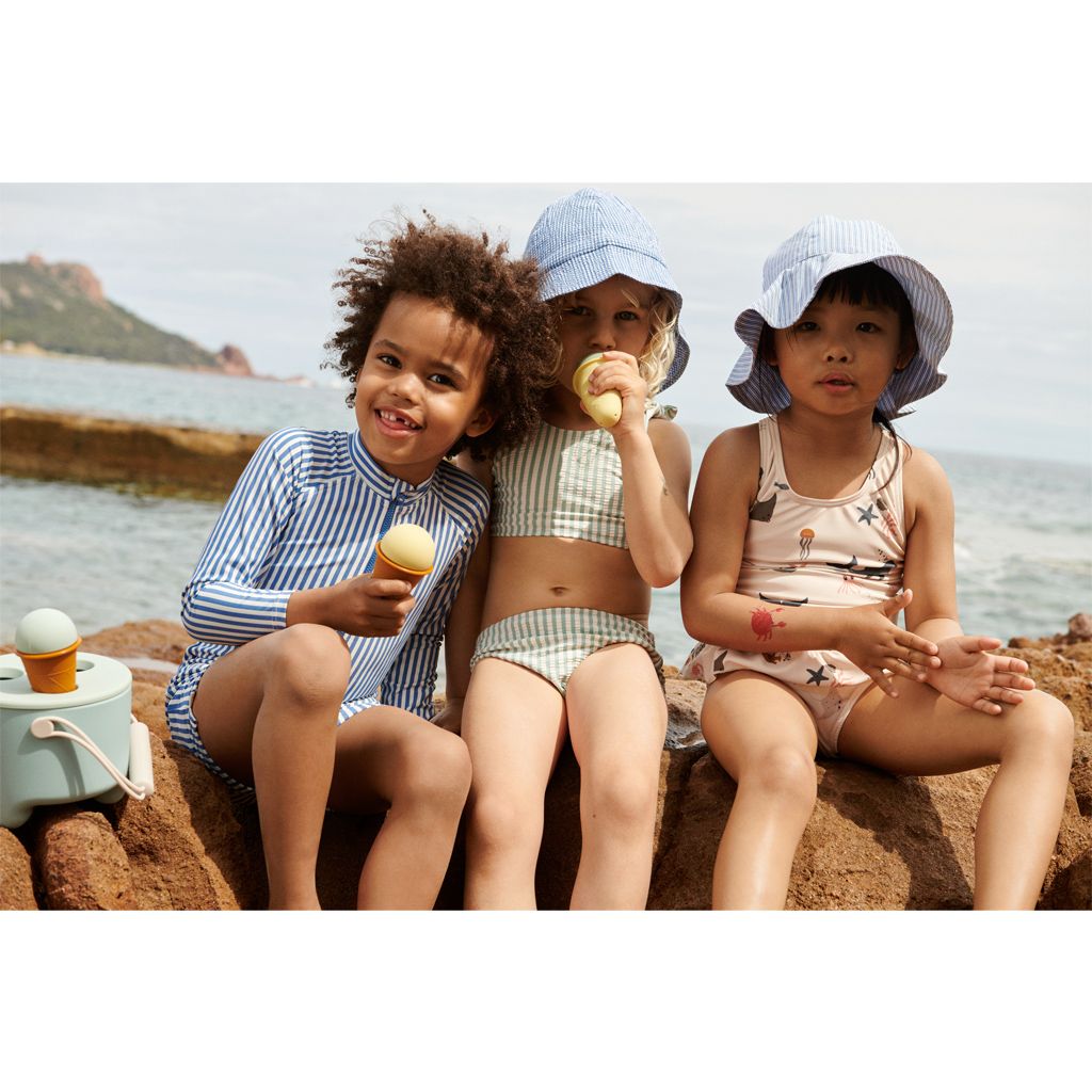 Três crianças na praia com roupa de banho e chapéus azuis, segurando gelados.