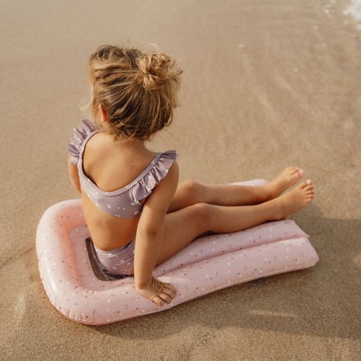Menina em fato de banho lilás sentada numa almofada insuflável rosa na areia