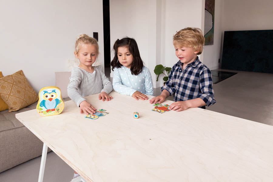 Três crianças a jogar jogo de tabuleiro numa mesa de madeira clara, caixa com desenho de coruja colorida visível.