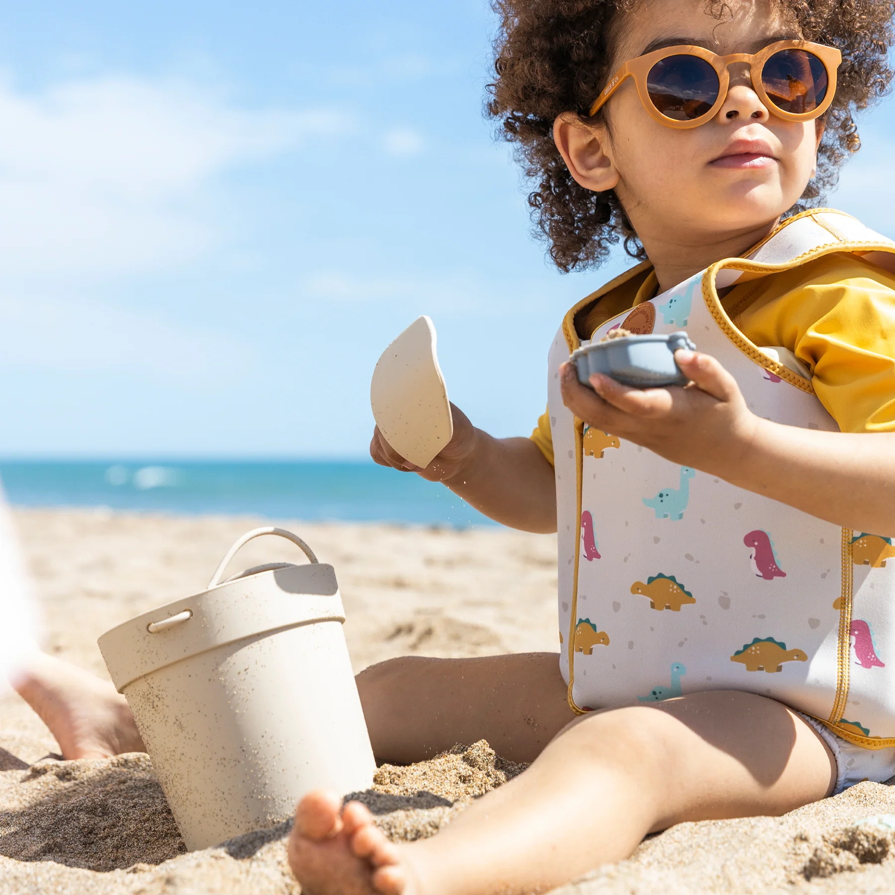 Criança na praia com colete salva-vidas e óculos de sol, brinquedos de praia ao redor
