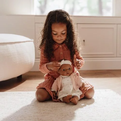 menina pequena com vestido rosa a brincar com boneca vestida de branco e rosa num quarto claro
