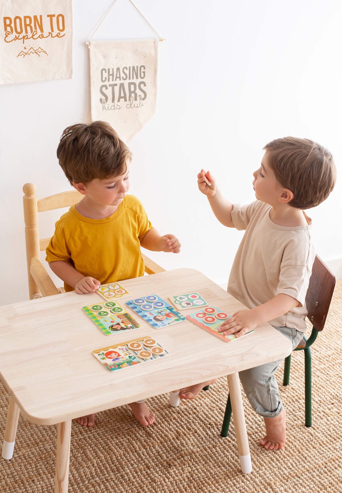 Duas crianças a jogar em mesa de madeira clara com cartelas coloridas e paredes com decoração em pano