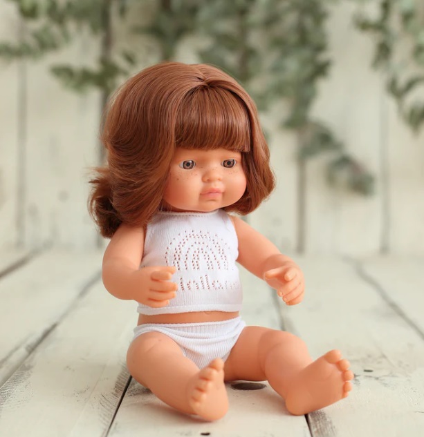 Boneca bebé com cabelo castanho e roupa branca sentada em chão de madeira