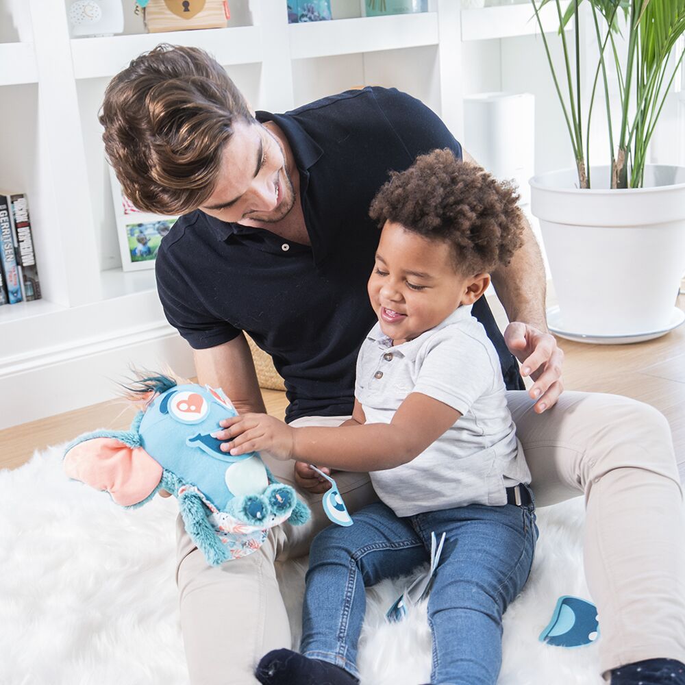 Criança a segurar brinquedo de pelúcia azul e rosa no colo de adulto num ambiente interior