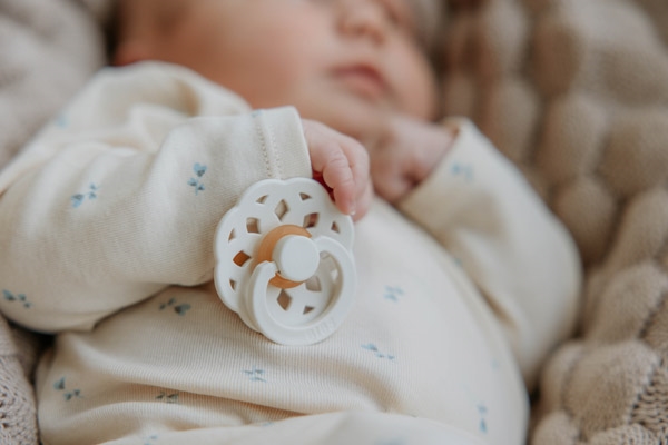 Bebé vestido com body branco com padrões azuis segurando chupeta creme numa manta de malha bege