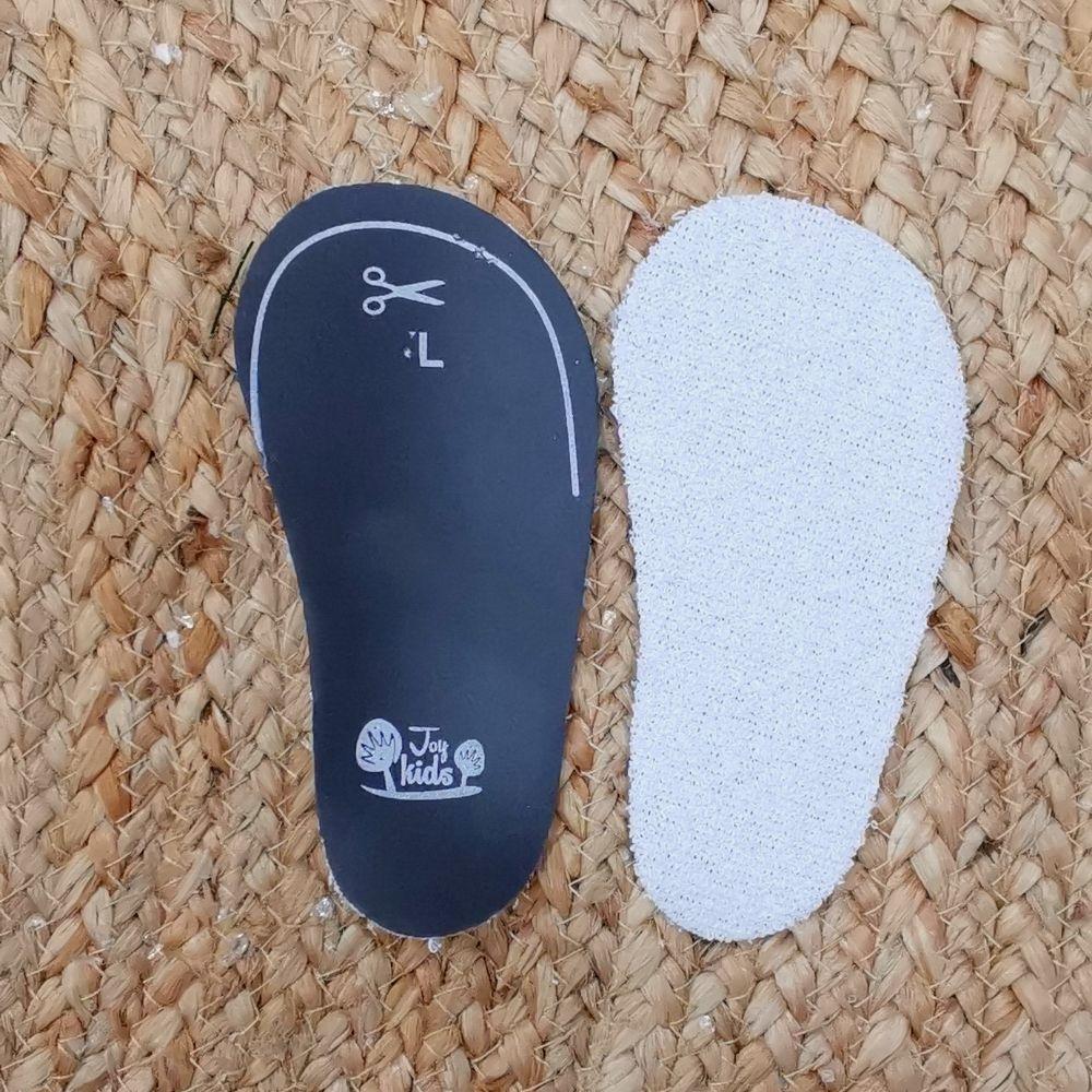 Palmilhas para crianças, preta com simbolo de tesoura e L, e branca lado a lado sobre superficie têxtil trançada