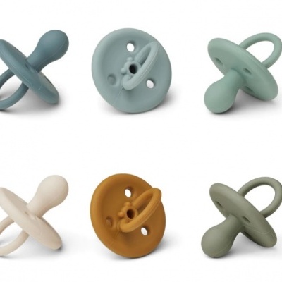 Seis chupetas de silicone em várias cores diferentes com anel, sobre fundo branco.