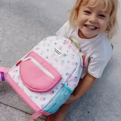 Criança sorridente com mochila branca com padrão de morangos e bolsos coloridos sentada no chão
