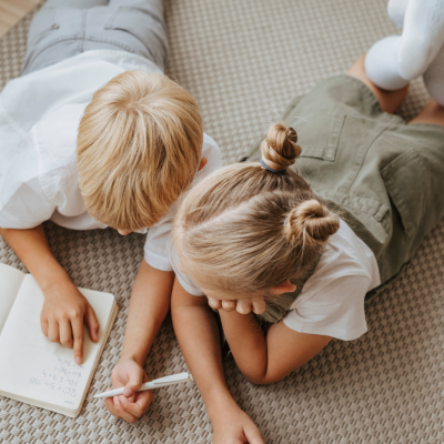 Duas crianças loiras deitadas numa carpete bege com um caderno e caneta.