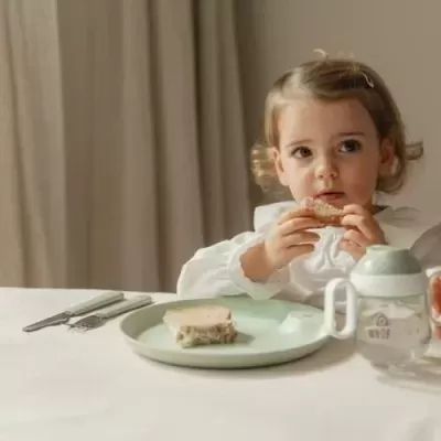 Criança pequena com camisa branca segurando pão à mesa branca com prato, talheres e copos