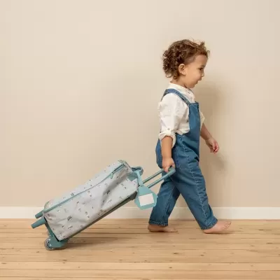 Criança puxando mala com padrão, vestindo jardineira azul, chão madeira clara e parede bege