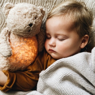 Criança dormindo com um urso de pelúcia iluminado e manta cinza texturada