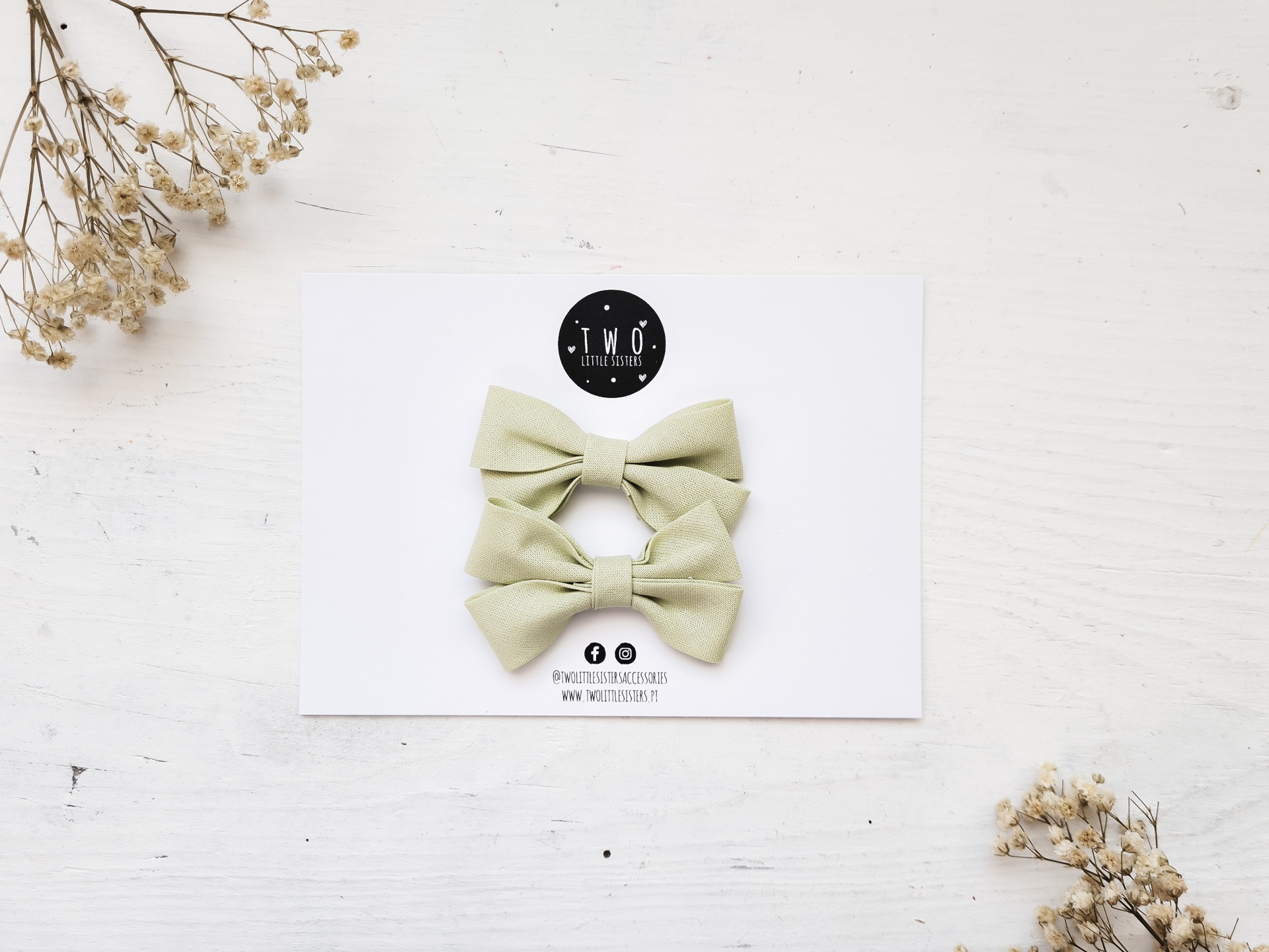 Dois laços de tecido verde claro numa tarjeta branca com logótipo TWO e flores secas ao redor