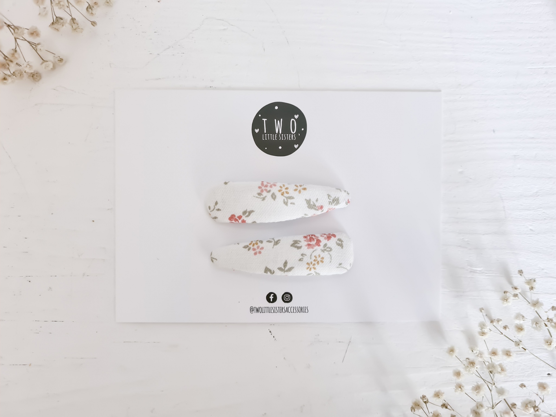 Molhas de cabelo branca com padrão floral em cartão branco com logotipo TWO LITTLE EXPLORERS e raminhos de flores secas