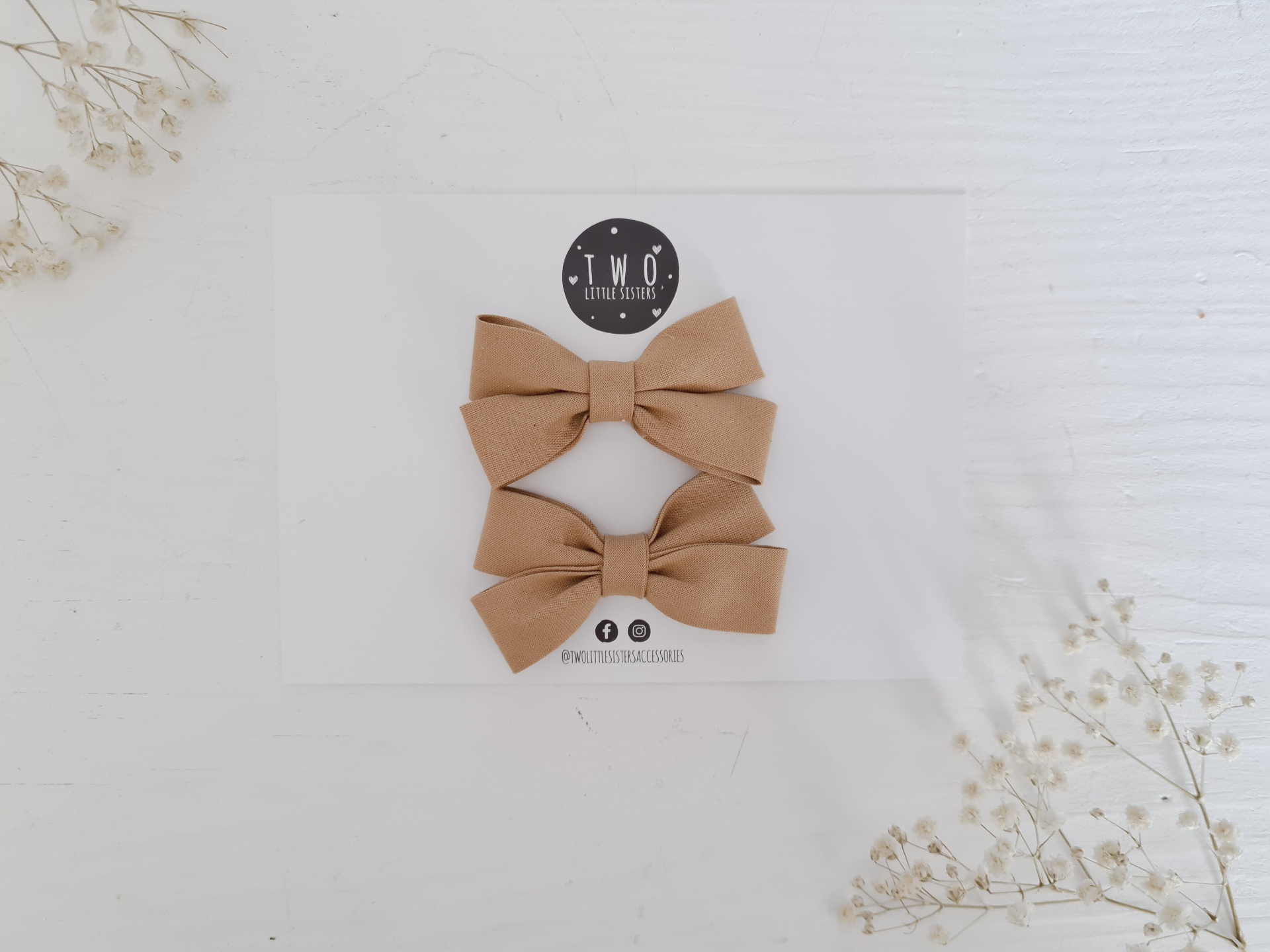 Dois laços para cabelo castanhos em tecido num cartão branco com logotipo T.W.O e flores secas