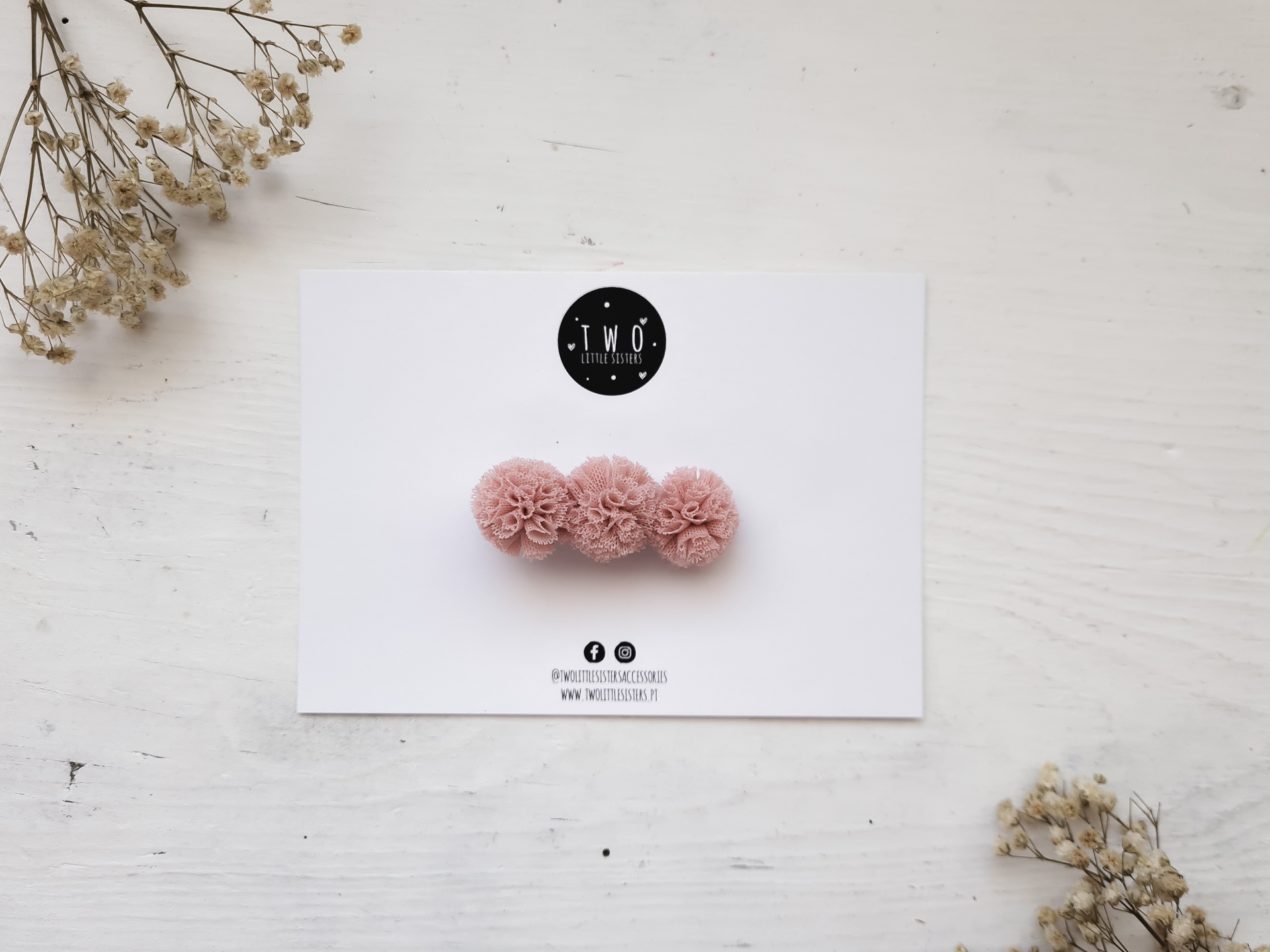 Clipe de cabelo com pompons rosa claro sobre cartão branco e fundo branco com flores secas.