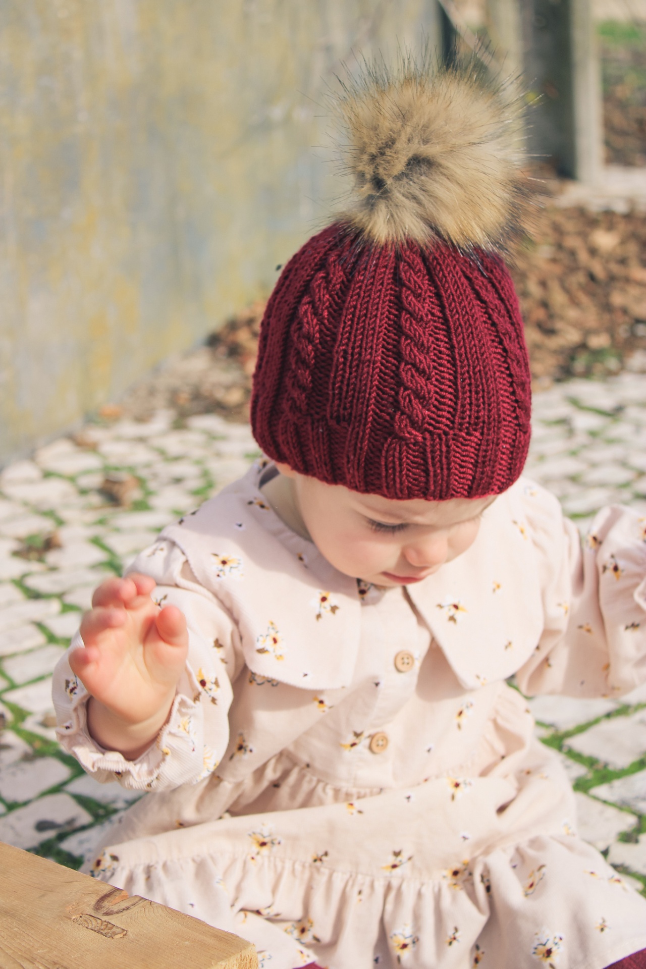 Bebé com gorro vermelho e pompom castanho claro e vestido creme floral sentado ao ar livre