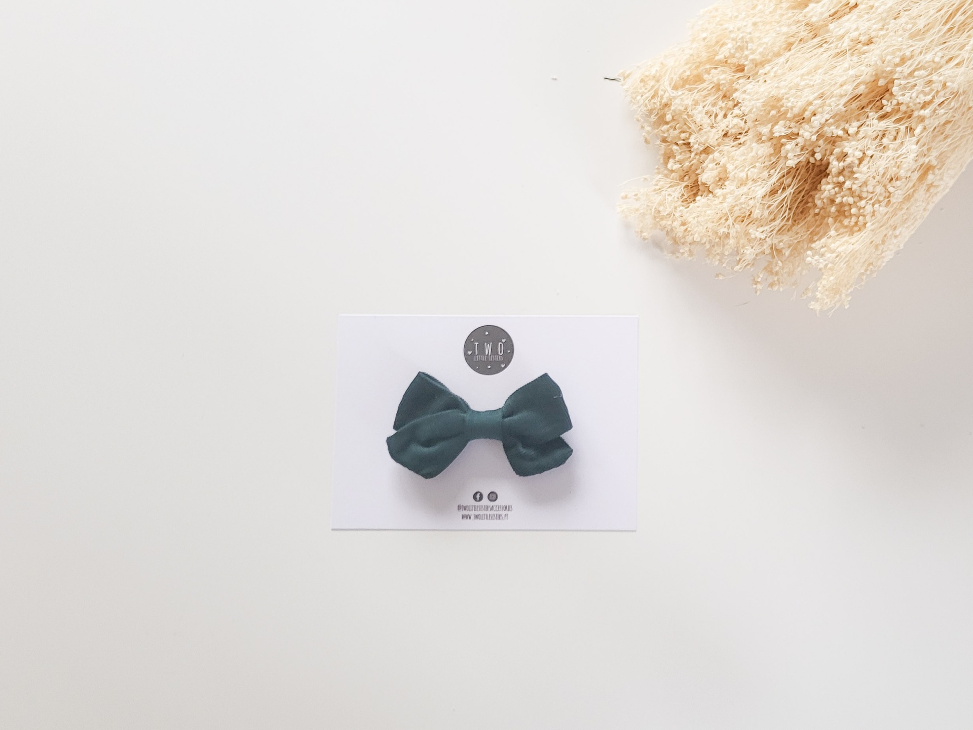 Laço para cabelo verde escuro em cartão branco com plantas secas ao lado