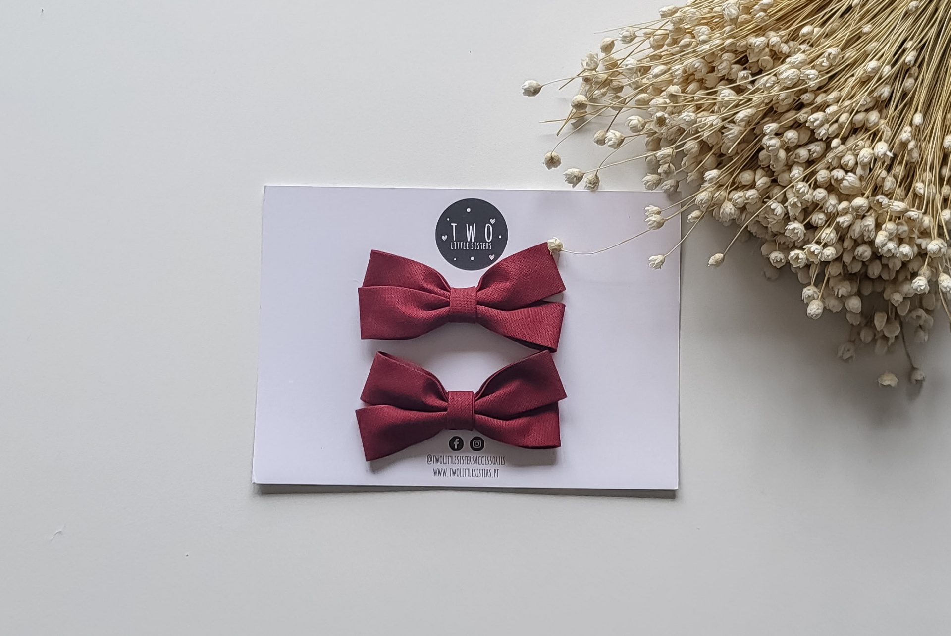 Dois laços bordô fixados num cartão branco com logótipo preto e branco ao lado de raminhos secos