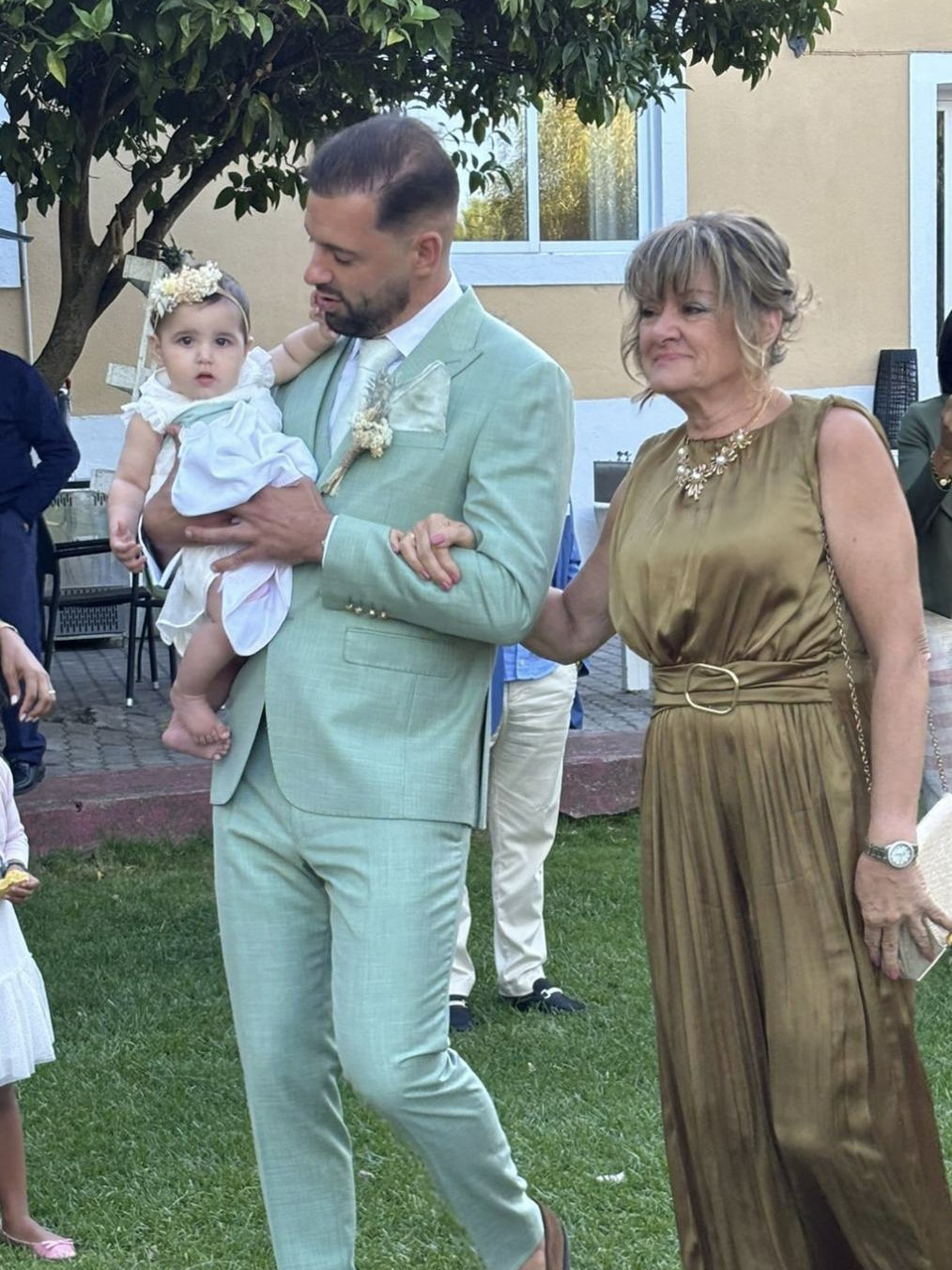 Homem de fato verde claro segura criança com vestido branco ao lado de mulher de vestido dourado em jardim