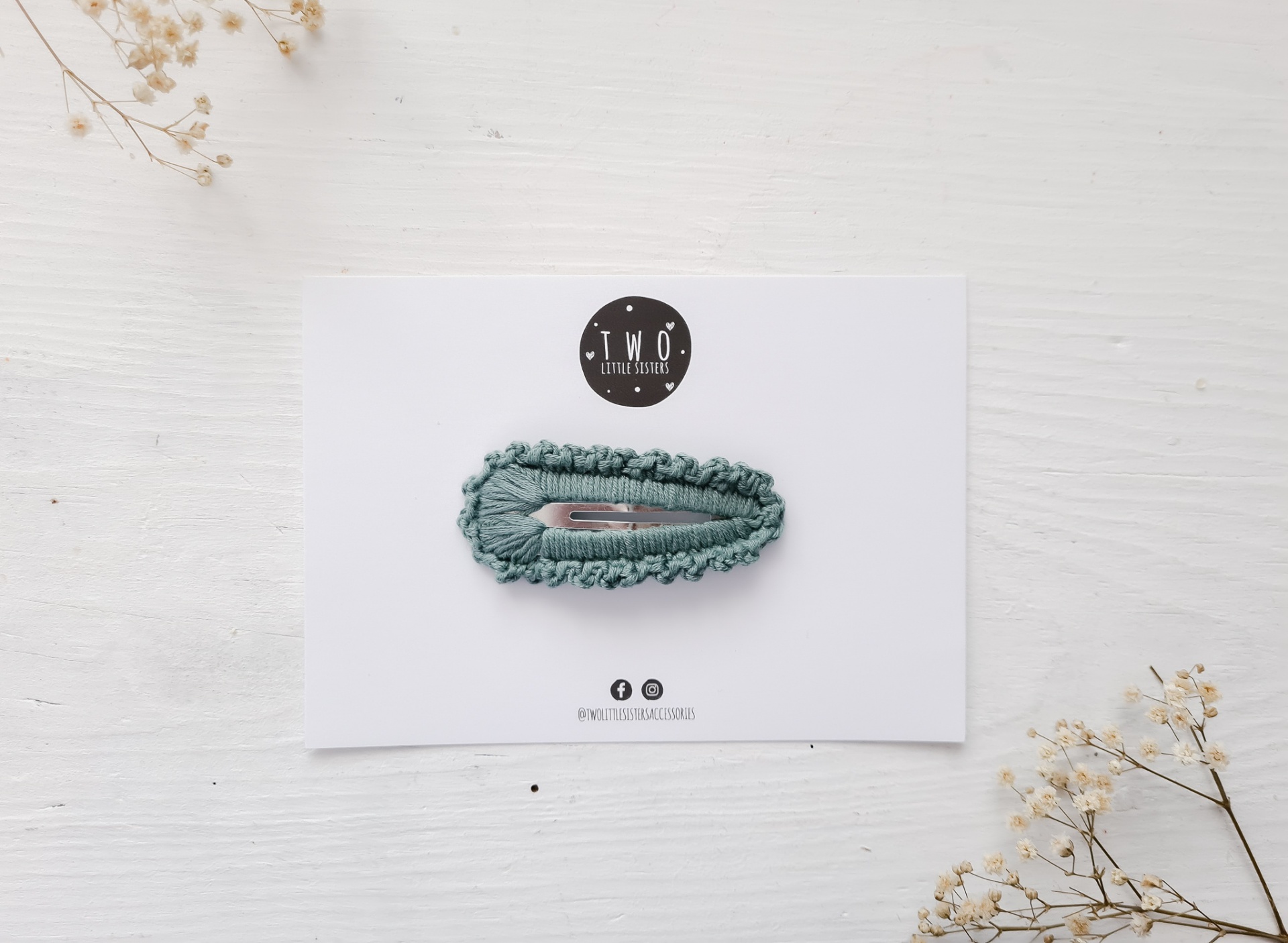 Grampo de cabelo verde em cartão branco com logo T.W.O e flores secas no fundo