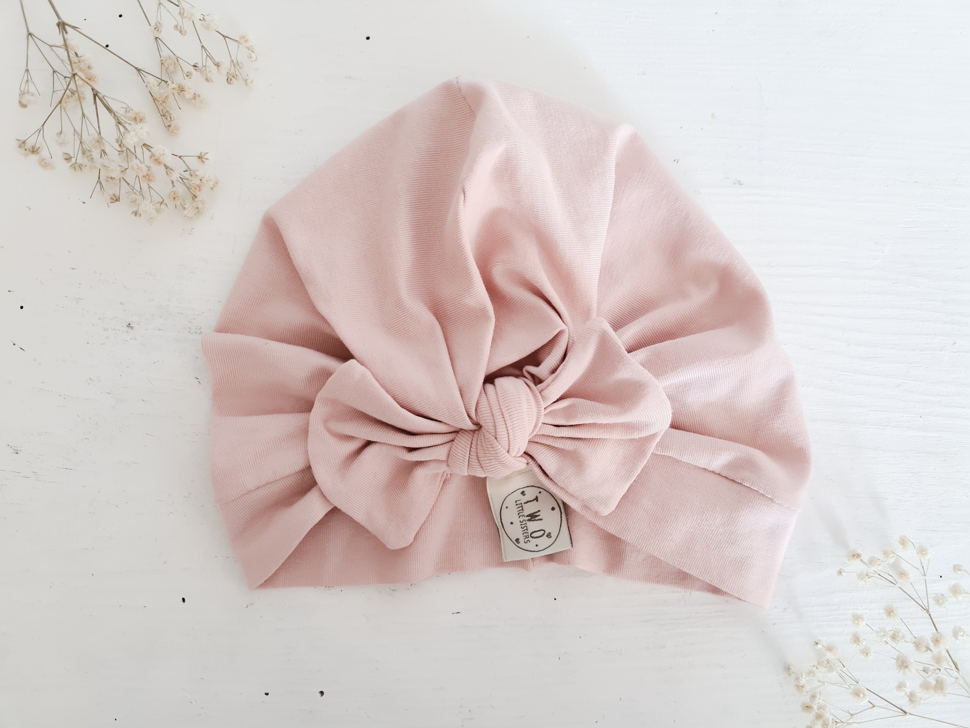 Touca cor-de-rosa claro com nó central sobre fundo branco e flores secas