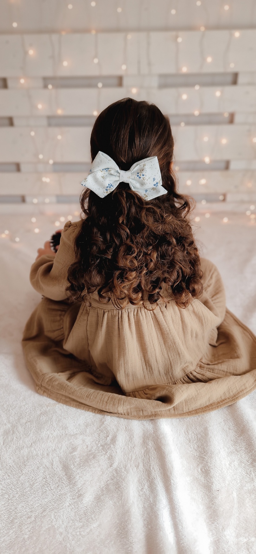 Menina com vestido castanho e laço branco no cabelo sentada de costas numa cama com luzes no fundo