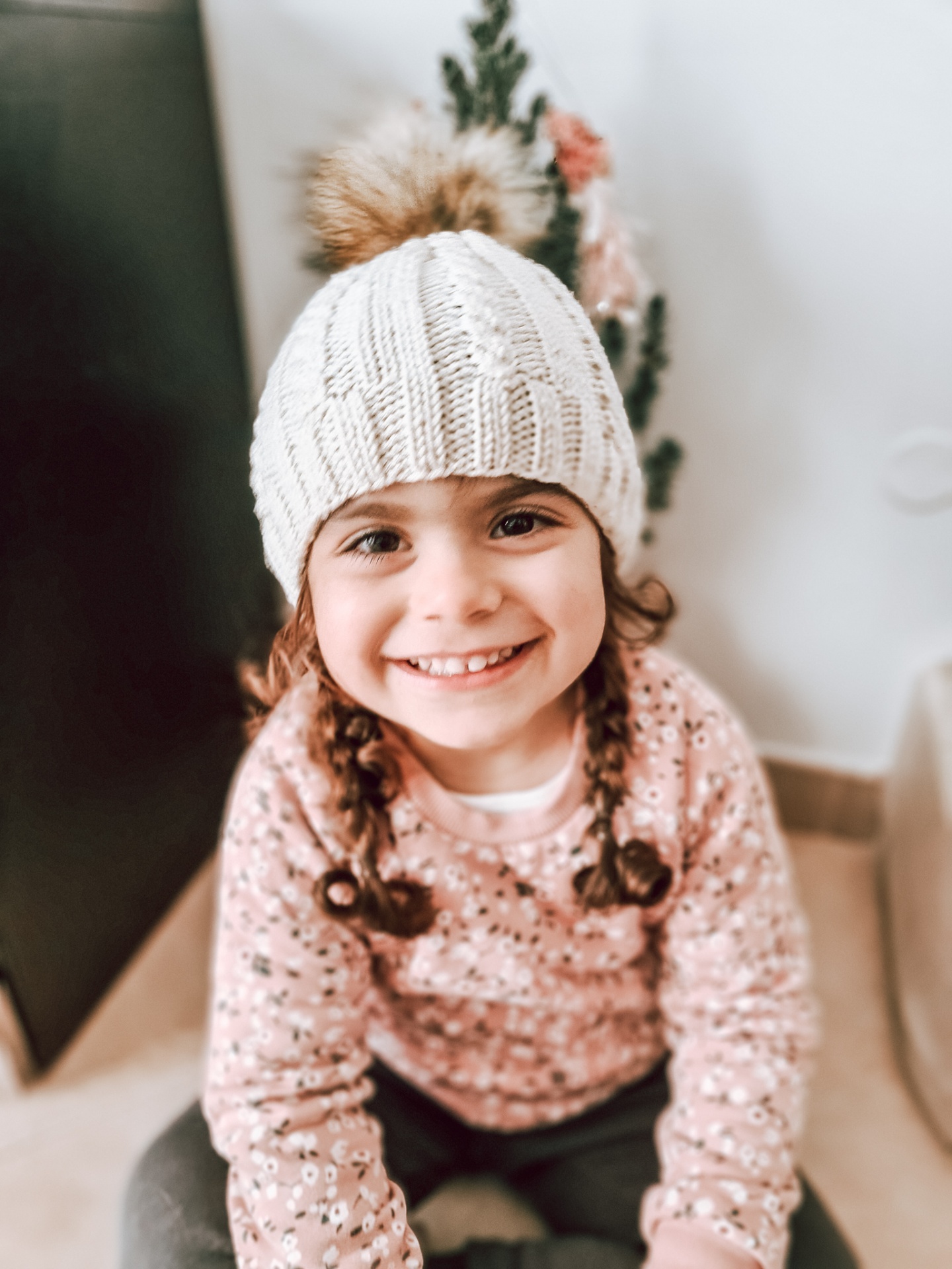 Criança com gorro branco de malha sentada e sorridente, com camisola rosa de flores