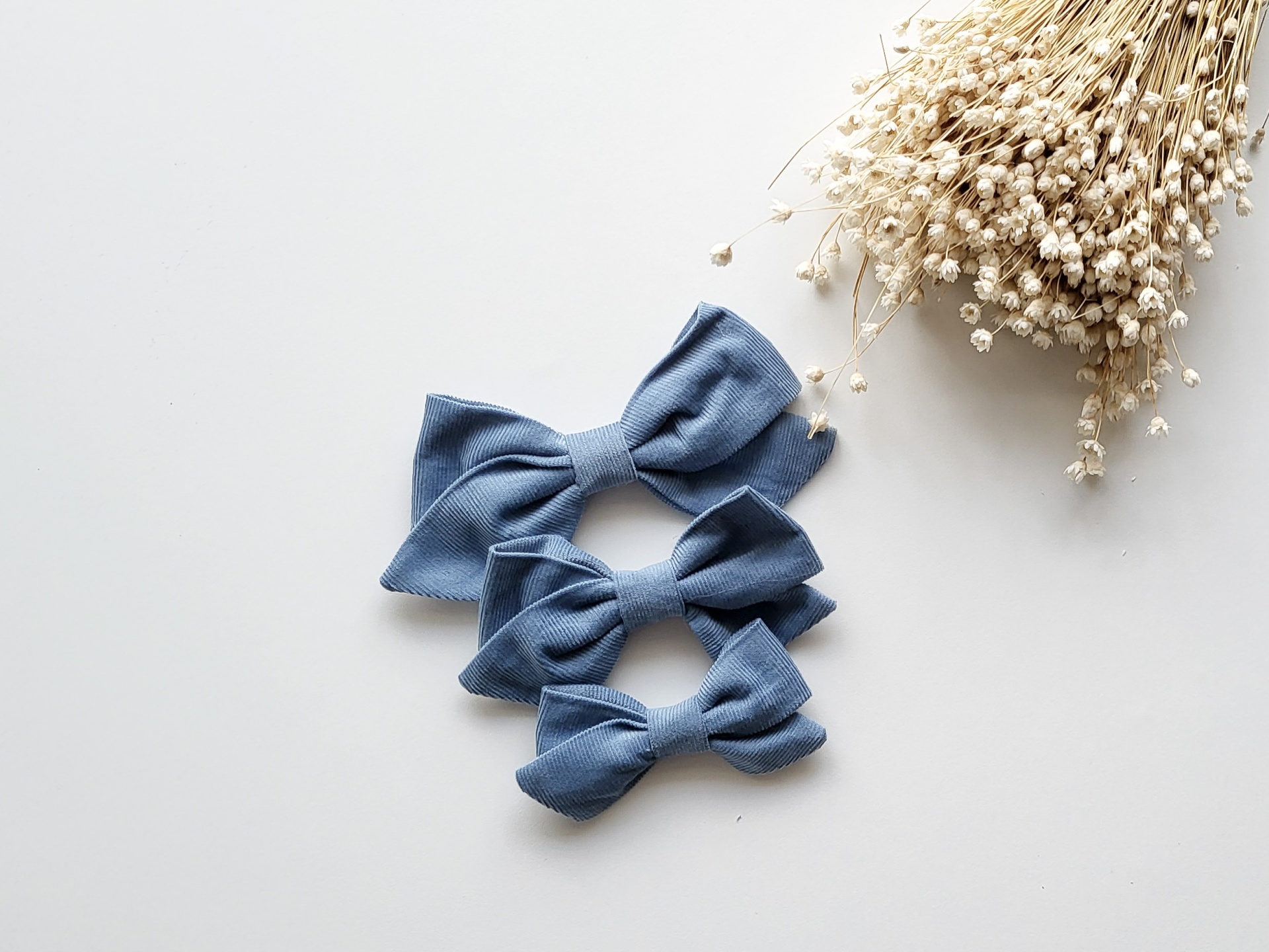 Três laços azuis em tecido sobre fundo branco com flores secas ao lado