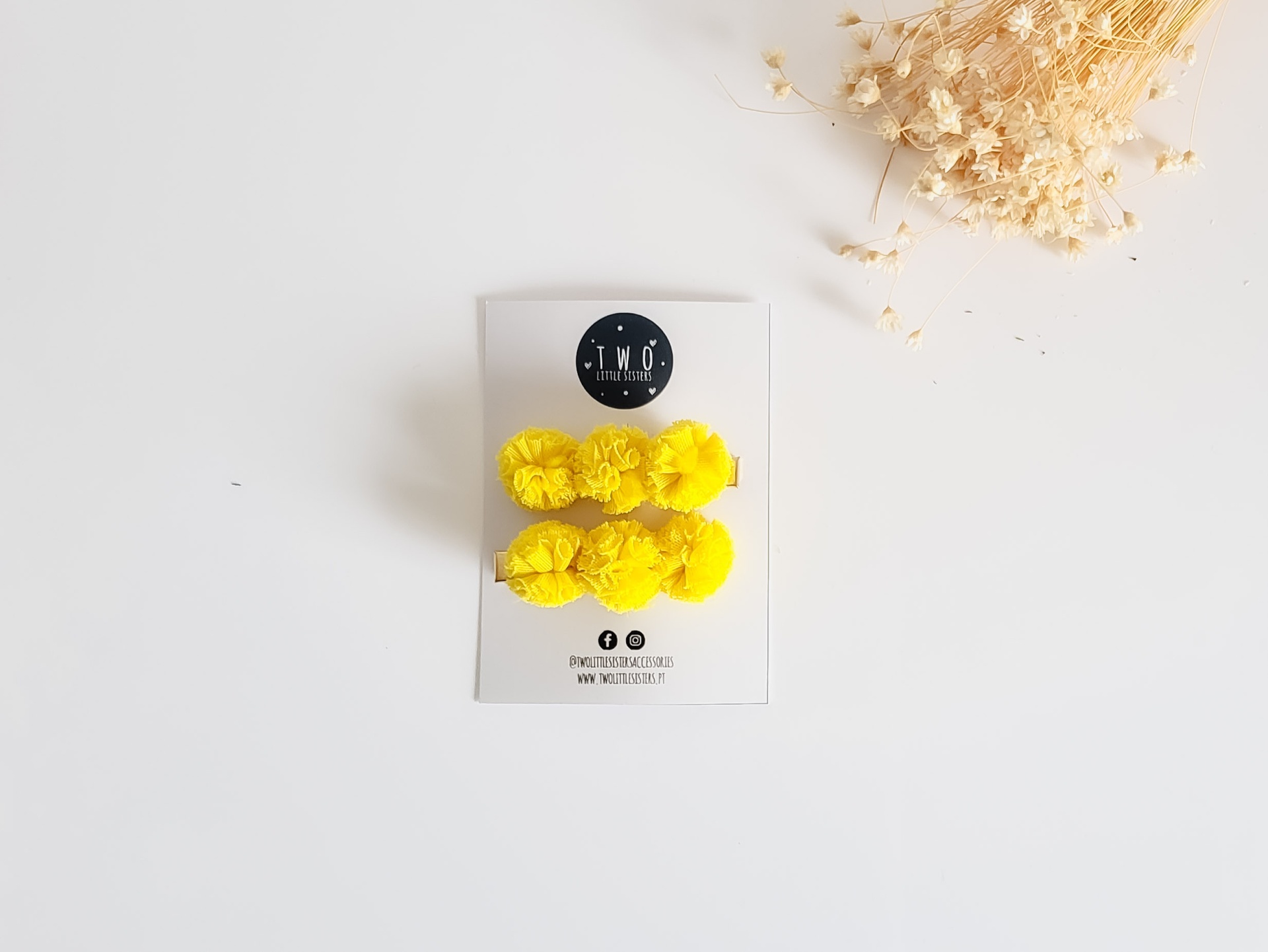 Brincos de pompons amarelos em cartolina branca com logotipo T.N.O e ramos secos ao lado