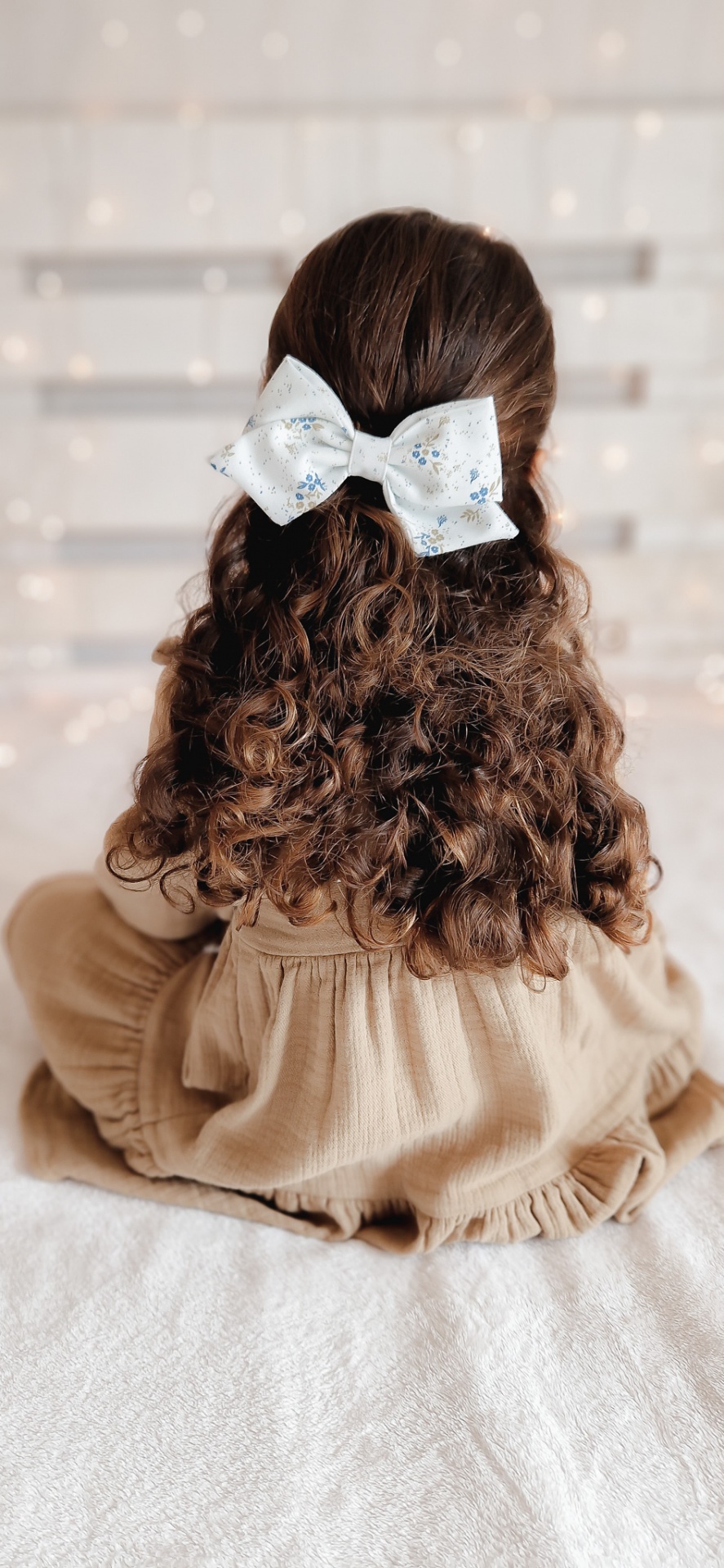 Criança com cabelo cacheado preso por laço branco decorado com flores, vestindo vestido bege sentada de costas.