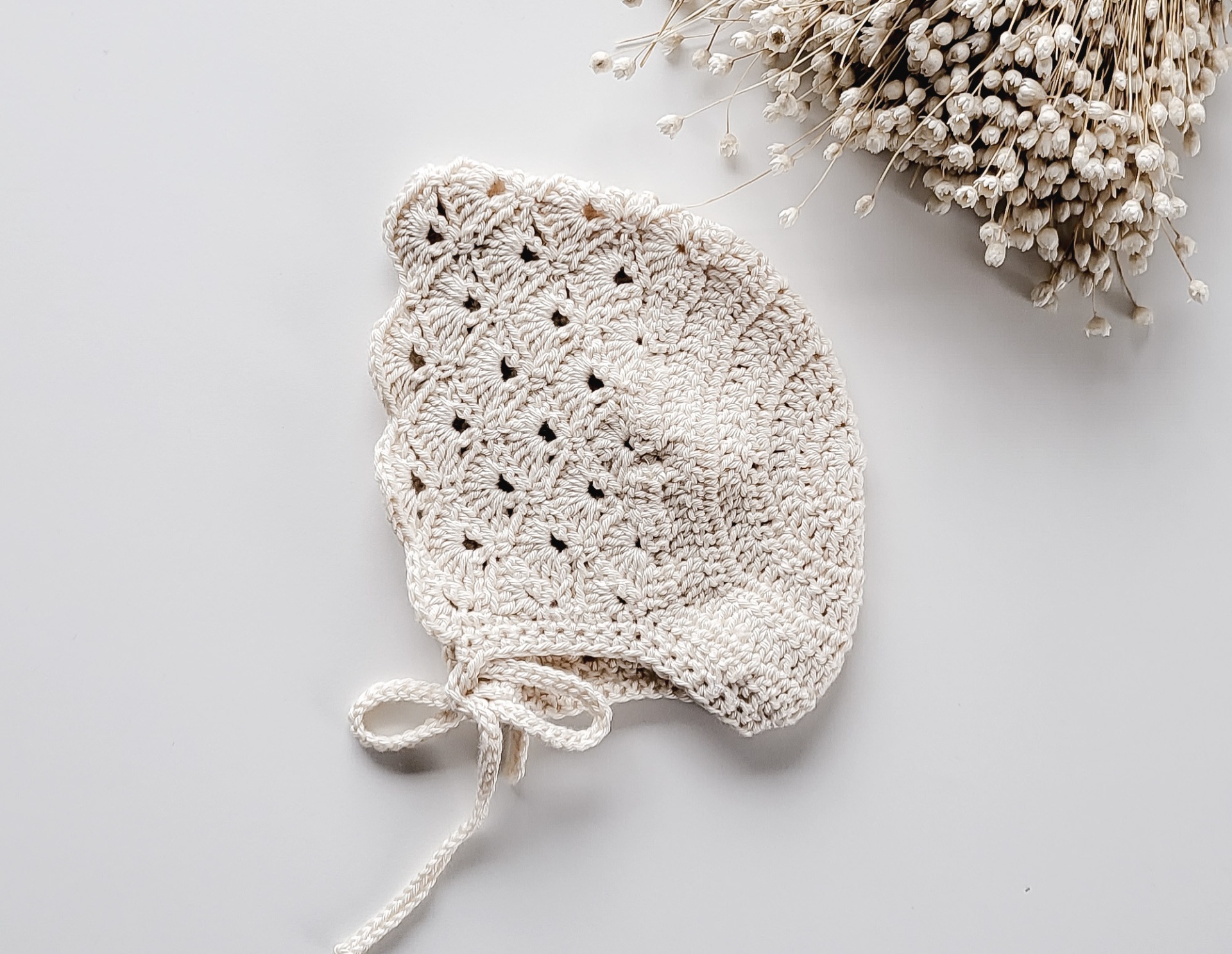 Gorro de malha cru com padrão vazado e fitas, ao lado de flores secas brancas.