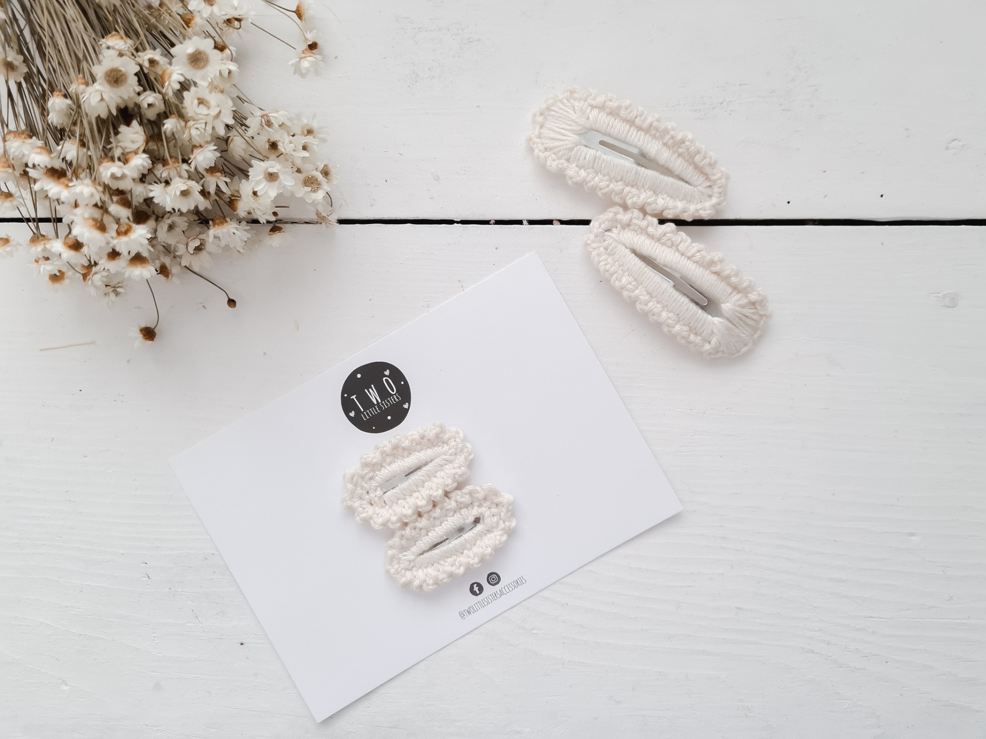 Três ganchos de cabelo brancos com textura de pérolas sobre fundo branco e cartão com texto e logotipos, ao lado de flores secas.