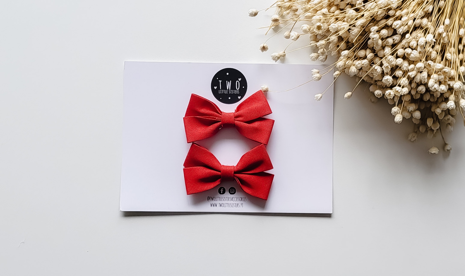Dois laços vermelhos em cartão branco com etiqueta T.N.O ACCESSORIES e flores secas ao lado