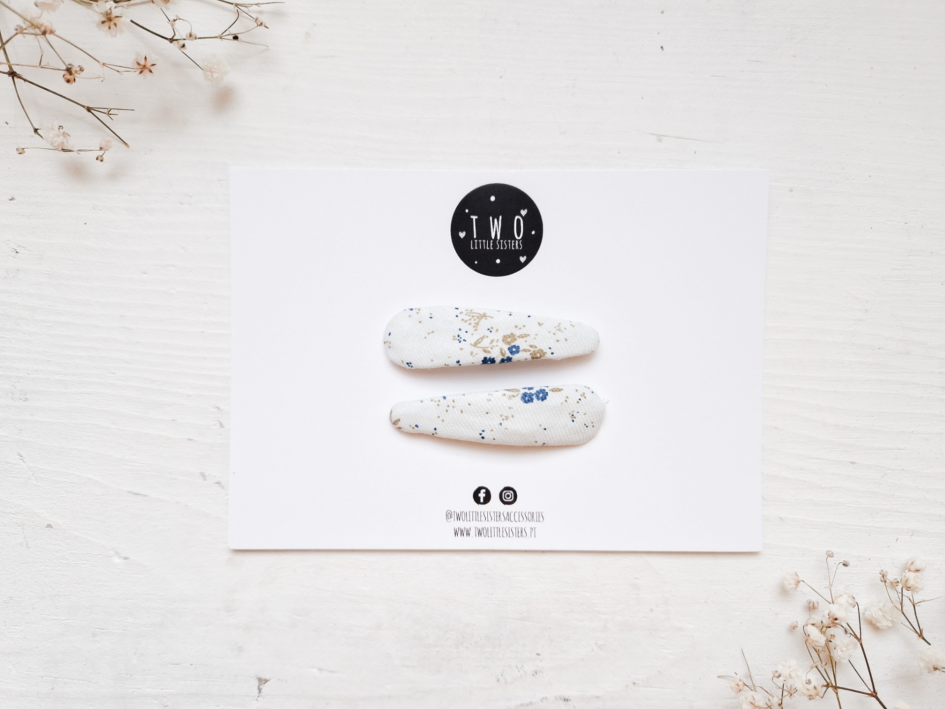 Presilhas de cabelo brancas com manchas azuis e beges sobre cartão branco com logótipo preto, em fundo branco com flores secas.