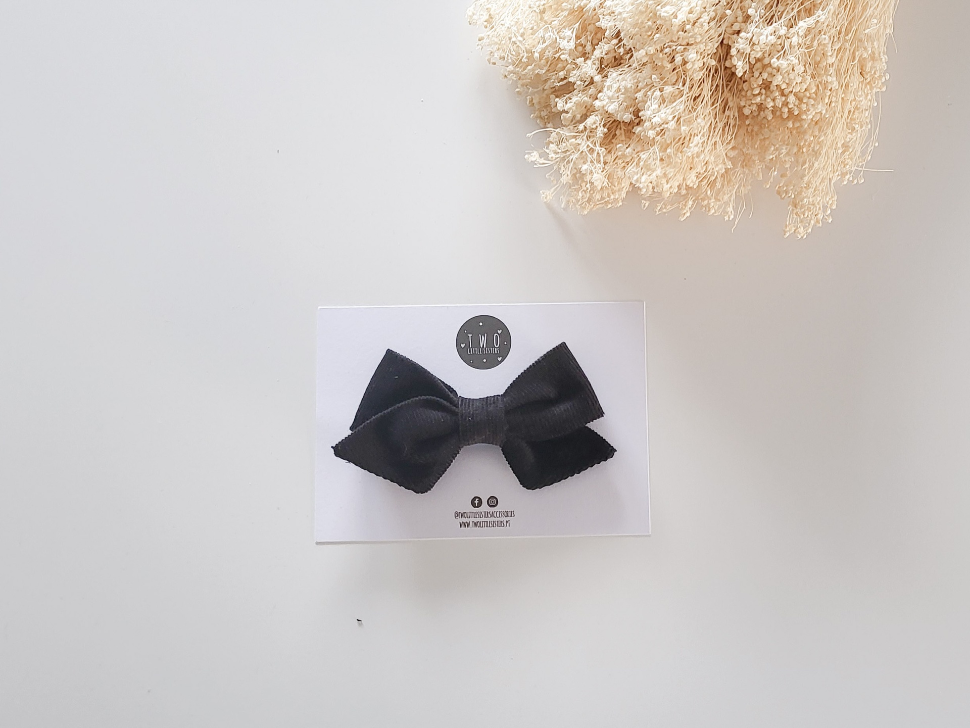 Laço de cabelo preto em cartão branco com texto à esquerda, raminho de flores secas ao lado