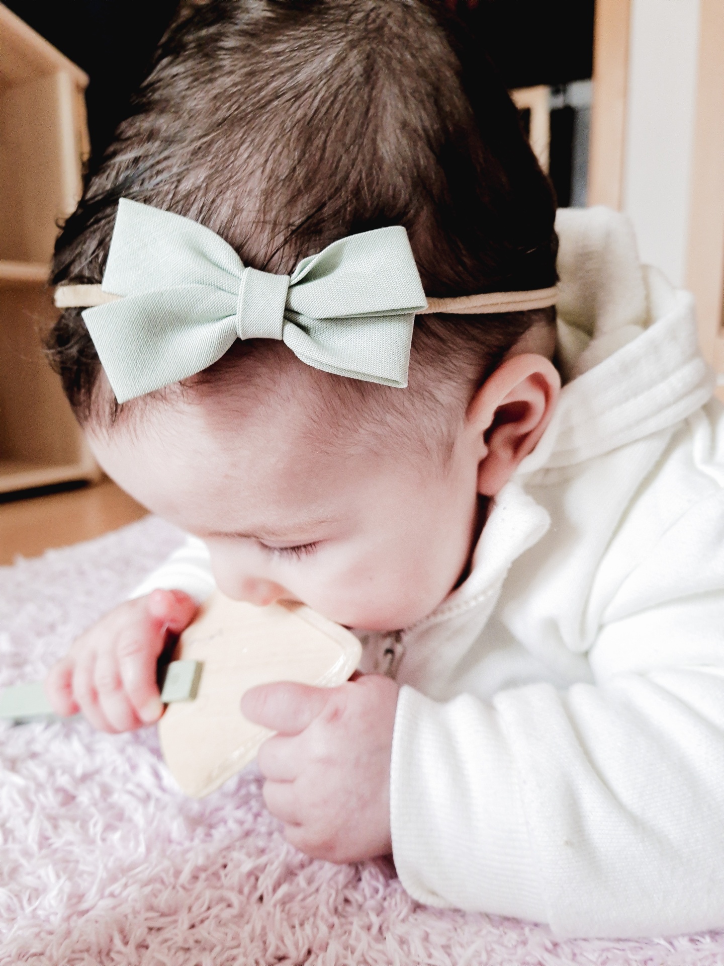 Bebé com faixa de cabelo laço verde claro e brinquedo de madeira sobre tapete rosa